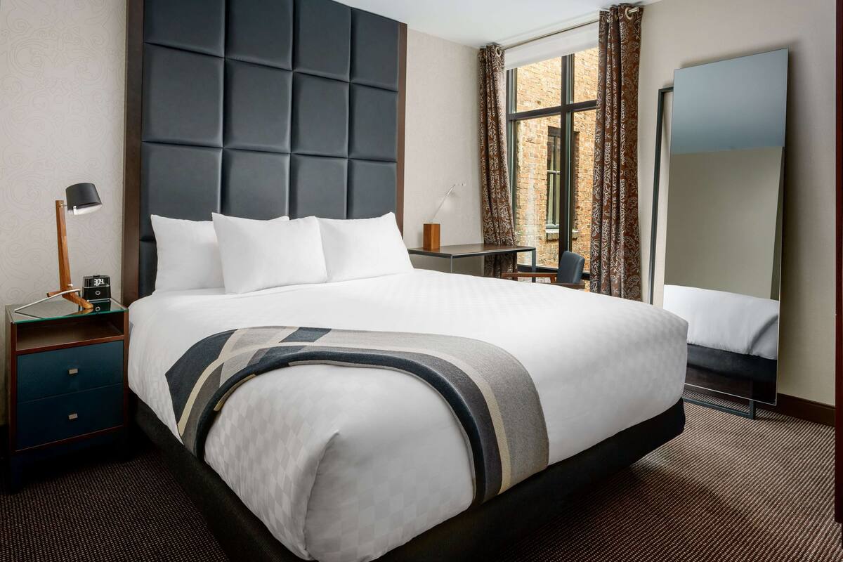 Ein Hotelzimmer mit einem großen Bett, einem Nachttisch, einem Schreibtisch, einem Sessel und einem großen Fenster mit Vorhängen.