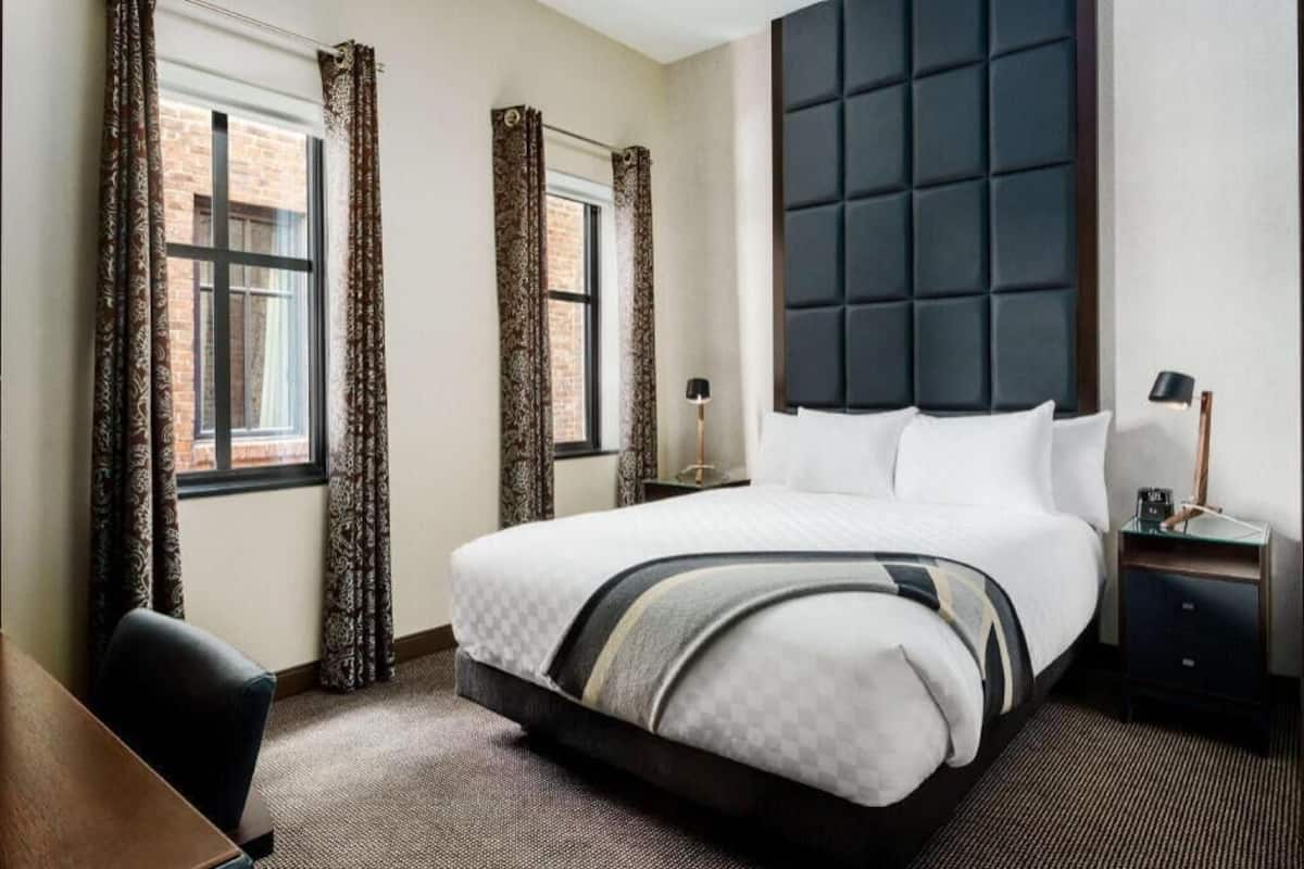 Ein Hotelzimmer mit einem großen Bett, einem Schreibtisch, zwei Fenstern mit Vorhängen, einem Kopfteil und einem Nachttisch mit Lampe.