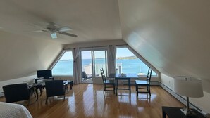 A spacious room with a wooden floor, a bed, a desk with a chair, a table with chairs, a ceiling fan, and a large window offering a view of the sea. - The Inn on the Wharf (Lubec)