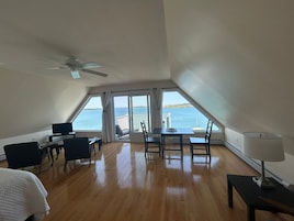 Amplia habitación con piso de madera, una cama, un escritorio con silla, una mesa con sillas, un ventilador de techo y un gran ventanal con vista al mar.