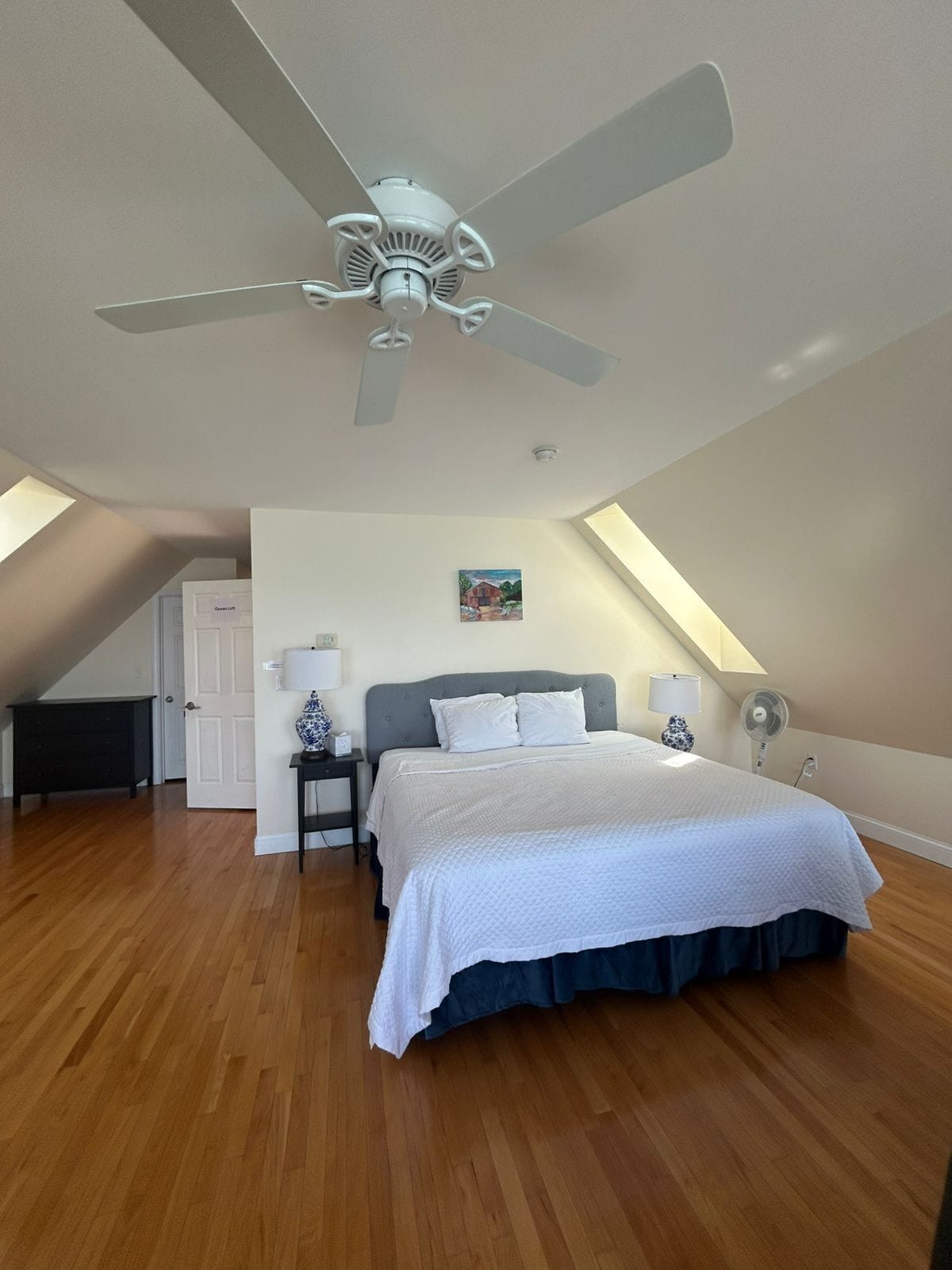 Ein Schlafzimmer mit einem Bett, einem Deckenventilator, einem Nachttisch, einer Lampe und einem Bild an der Wand.