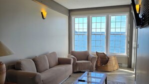 A living room with a beige sofa, a glass coffee table, and a view of the sea through large windows. - The Inn on the Wharf (Lubec)
