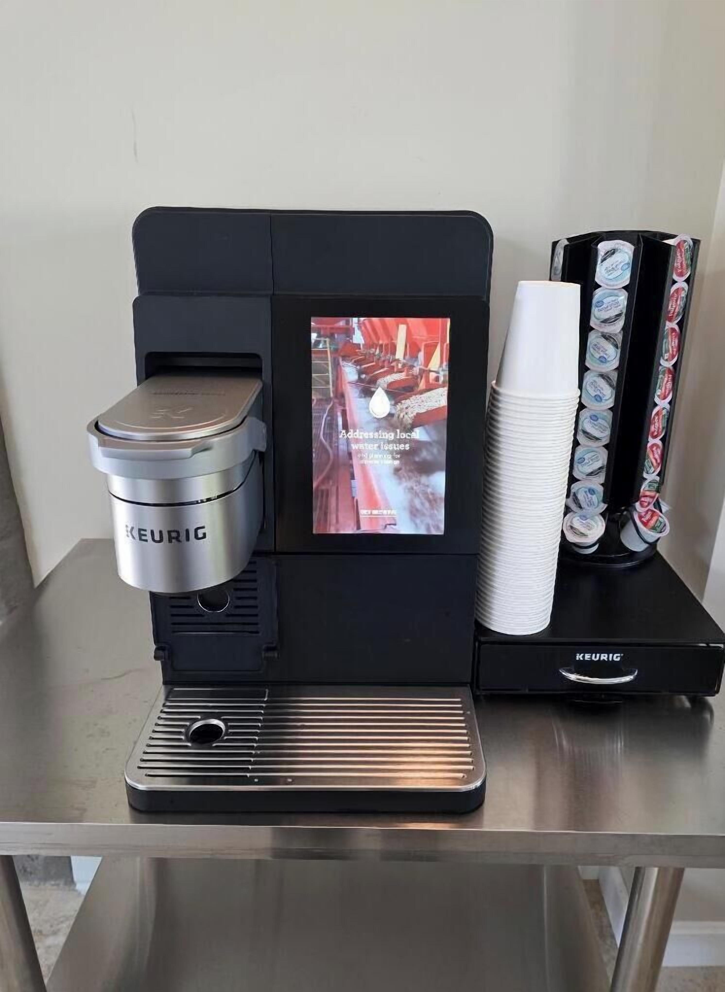 Eine Keurig-Kaffeemaschine mit digitalem Display, einem Kaffee-Pod-Halter und einem Stapel Einwegbecher auf einem Tisch.