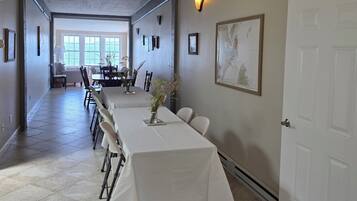 Un grand espace dédié à la salle à manger, avec une longue table, des chaises et un vase rempli de fleurs. Les murs sont ornés de tableaux encadrés et une porte se trouve sur le côté droit.