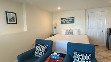 A hotel room with two blue armchairs, a small table with a red book, a bed with a 'welcome home' pillow, a fish-themed artwork, and a white door.
