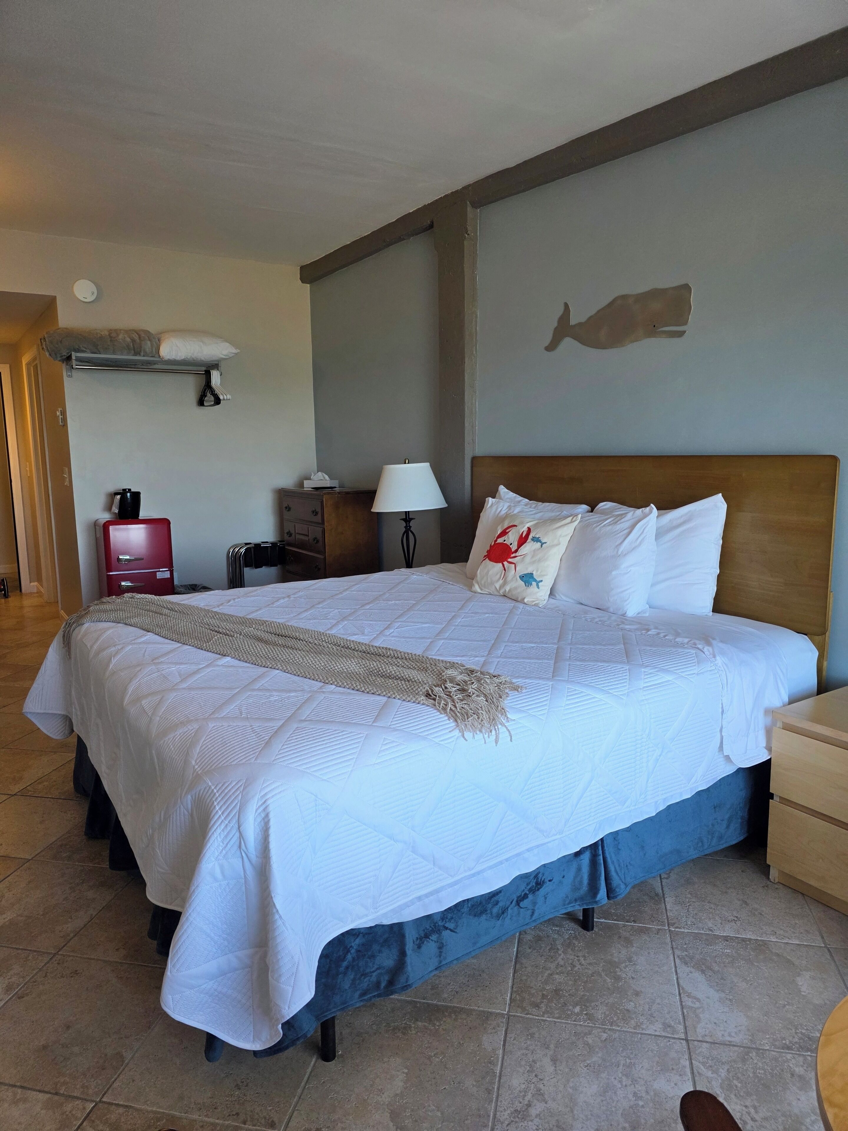 A hotel room with a large bed, a nightstand, a lamp, a red suitcase, a wall-mounted shelf, and a door.