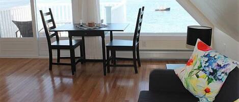 Habitació amb taula de dinar i cadires, un sofà negre amb coixí floral, i vista al mar a través de grans finestres.
