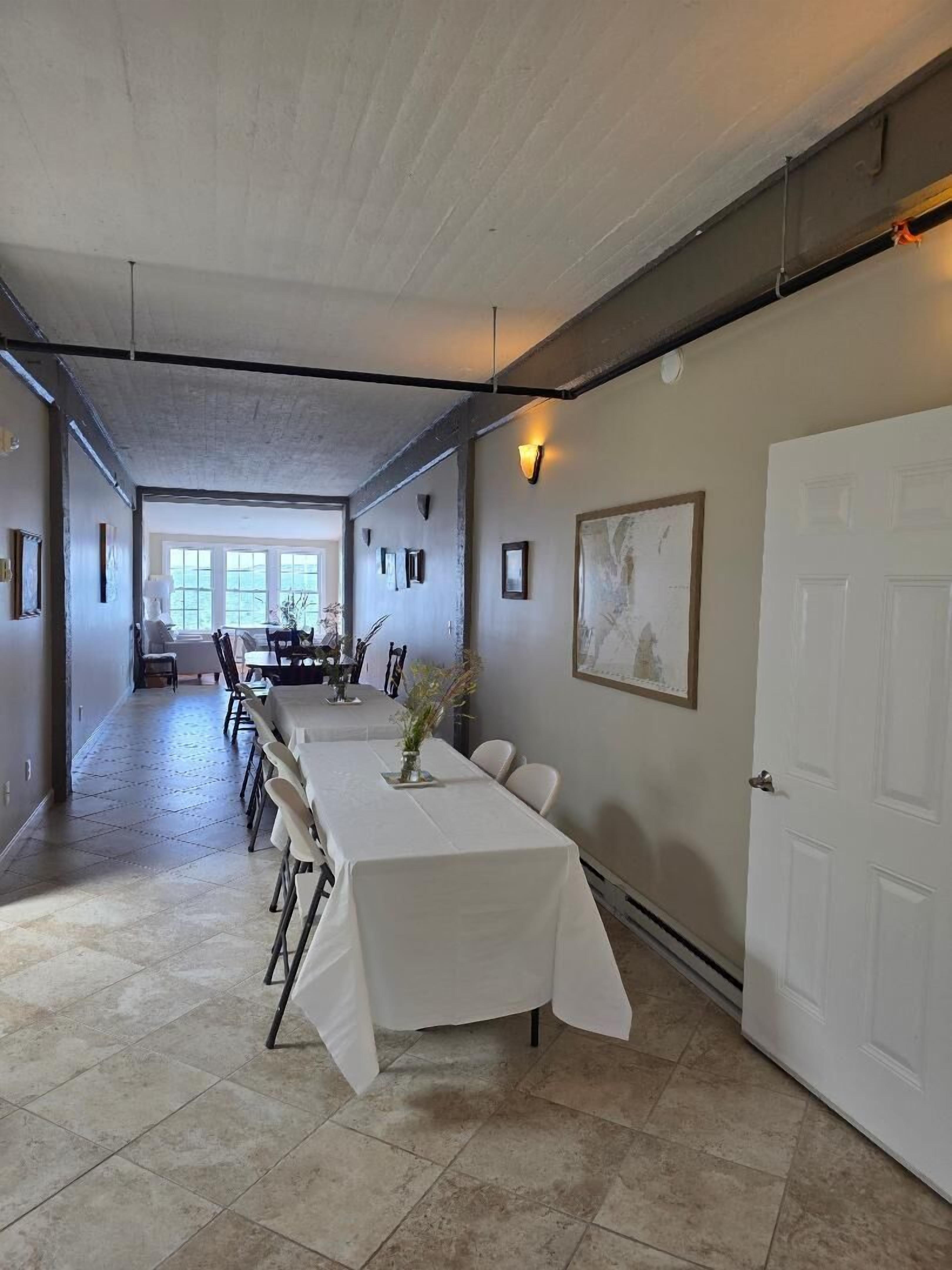 Une salle à manger spacieuse avec une longue table, des chaises et un vase de fleurs. Il y a une porte sur la droite et une fenêtre à l’arrière-plan.