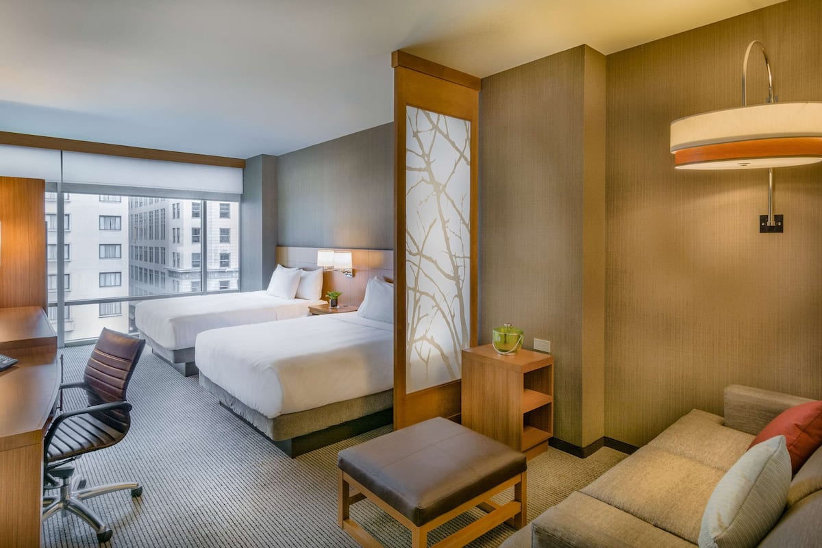 Une chambre d’hôtel avec deux lits, un bureau avec un téléphone, une chaise, un canapé, une petite table avec une plante et une grande fenêtre donnant sur la ville.