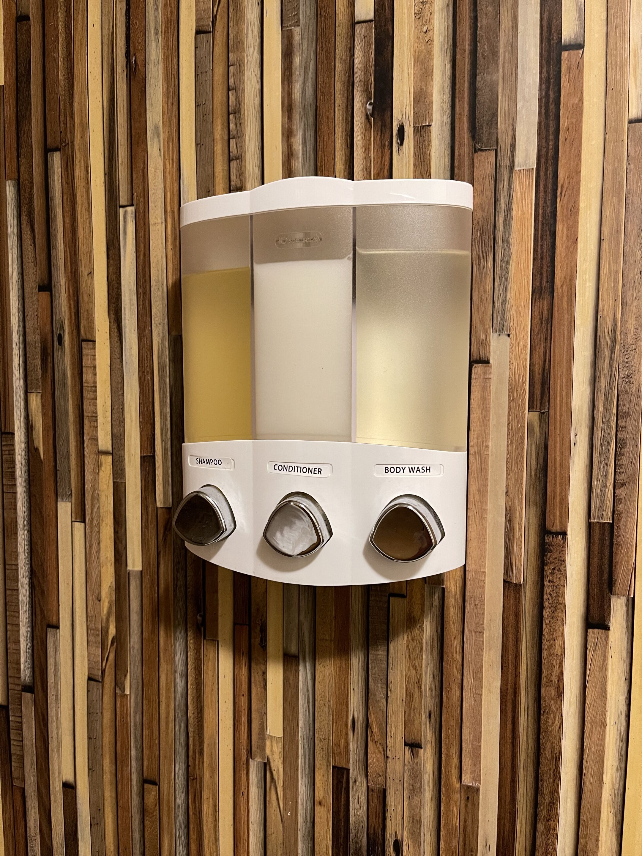 Dispensador de pared con tres compartimentos etiquetados para champú, acondicionador y gel de ducha.