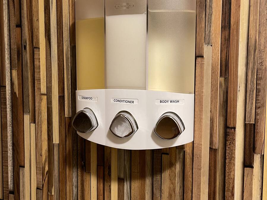 A wall-mounted dispenser with three compartments labeled for shampoo, conditioner, and body wash.