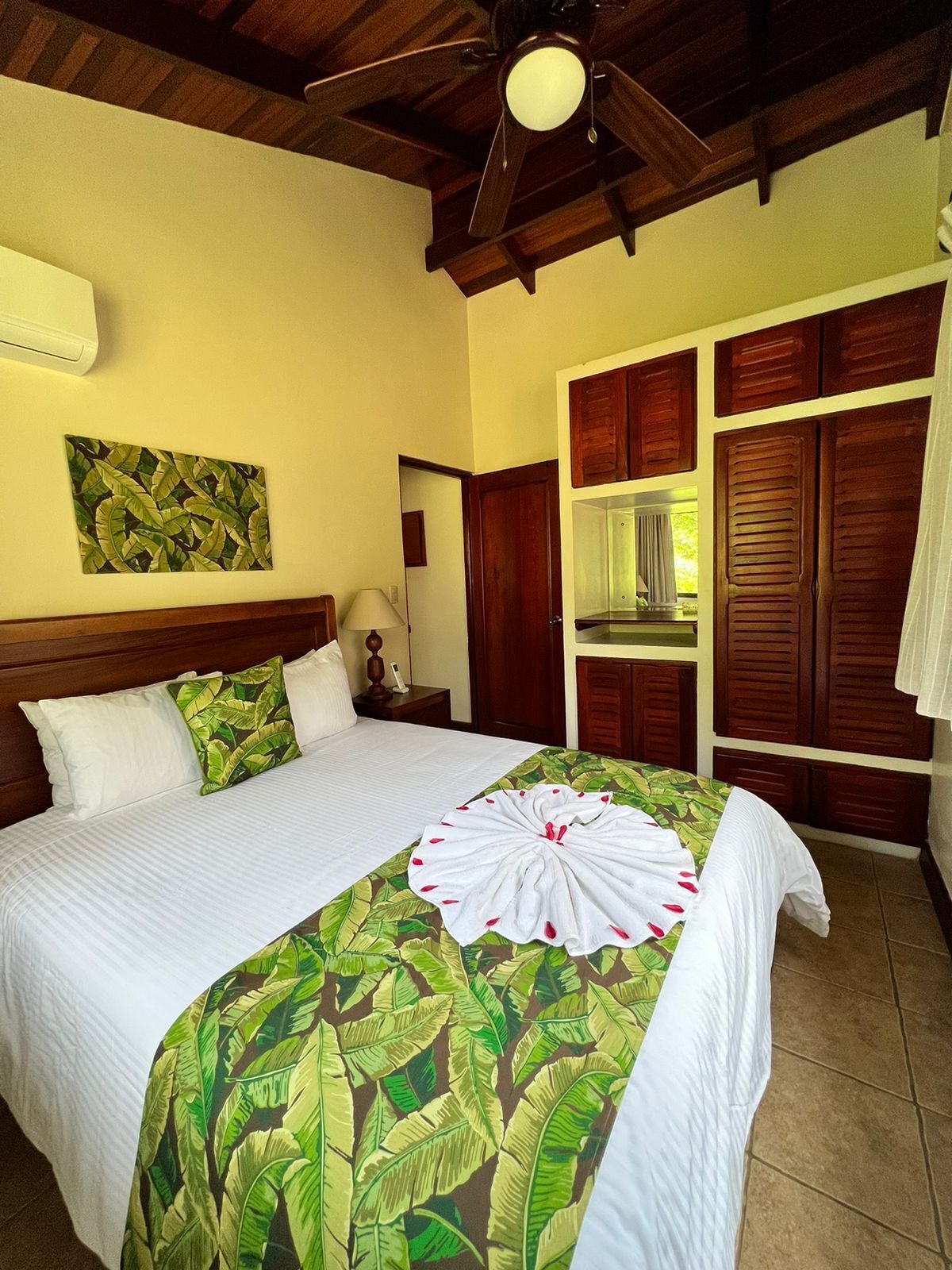 Une chambre d’hôtel avec un grand lit, un ventilateur de plafond, une fenêtre avec des volets en bois et un tableau au mur.