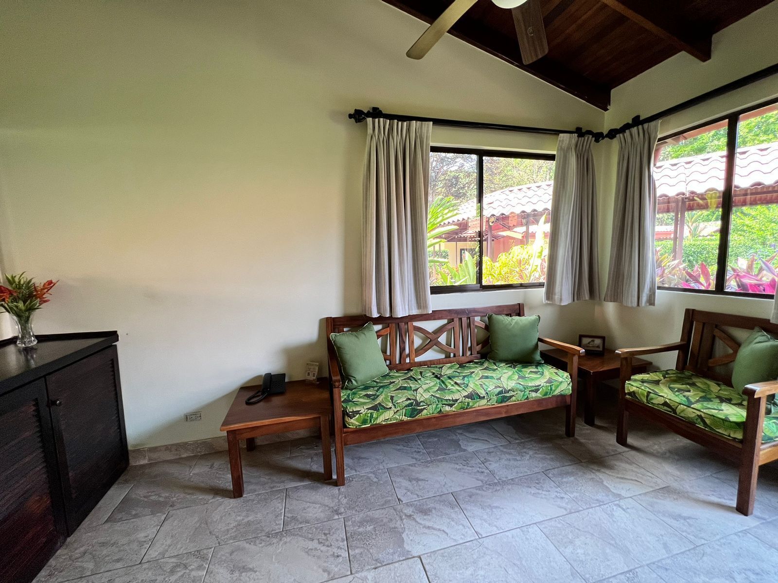 Una habitación con sofá y mesa de madera, además de una silla. Hay ventanas con cortinas, un ventilador de techo y vistas a zonas verdes.