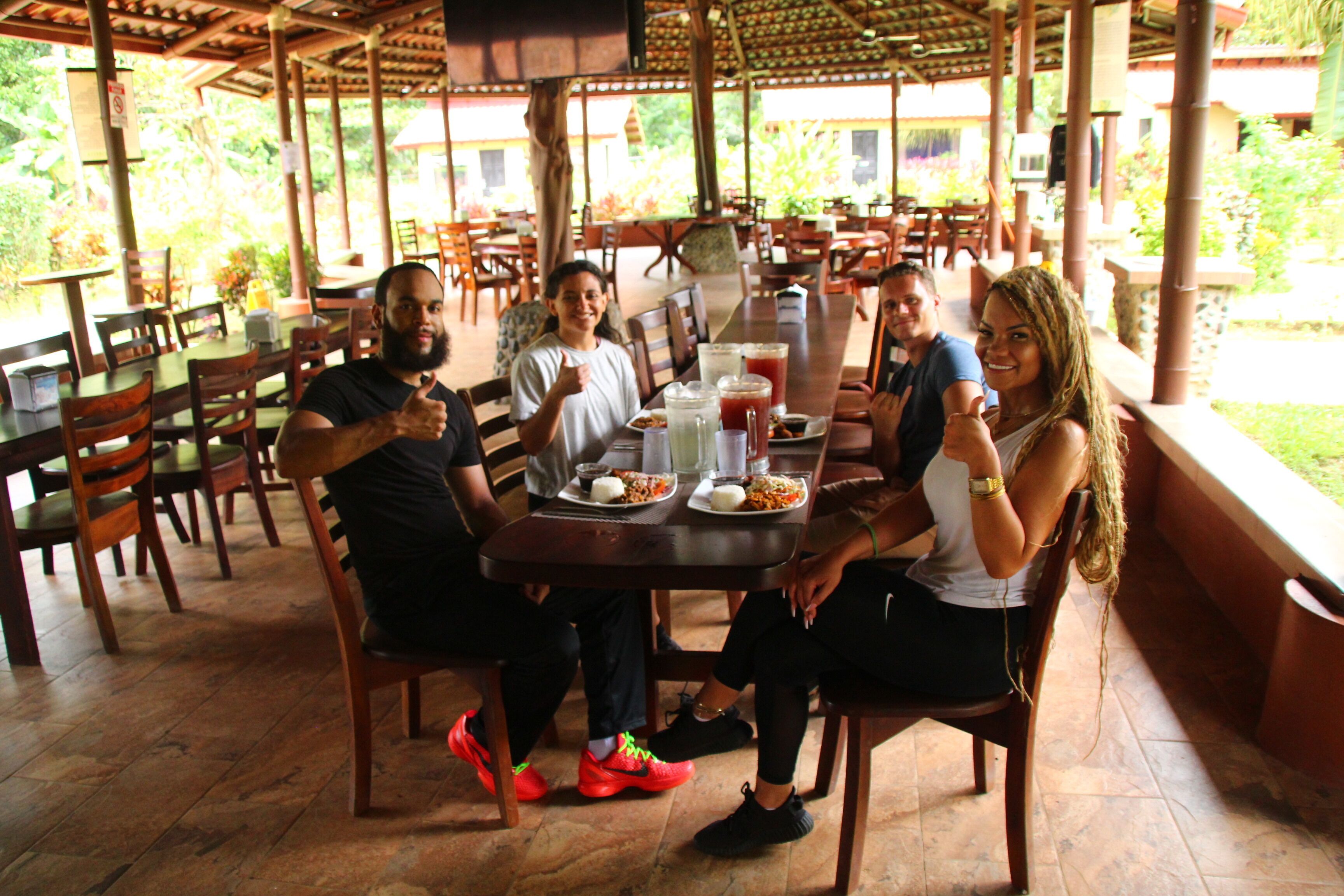 Un grupo de personas sentadas alrededor de una mesa con comida y bebidas, en un espacio de comedor al aire libre con muebles de madera y techo cubierto.
