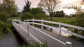 A wooden bridge over a calm water body, surrounded by greenery and a distant building. - Pheasant Field B&B (Carlisle)