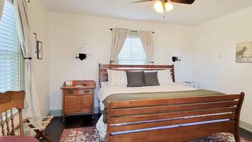 A bedroom with a wooden bed, a nightstand, a desk, a chair, a patterned rug, a ceiling fan, and a window with curtains.