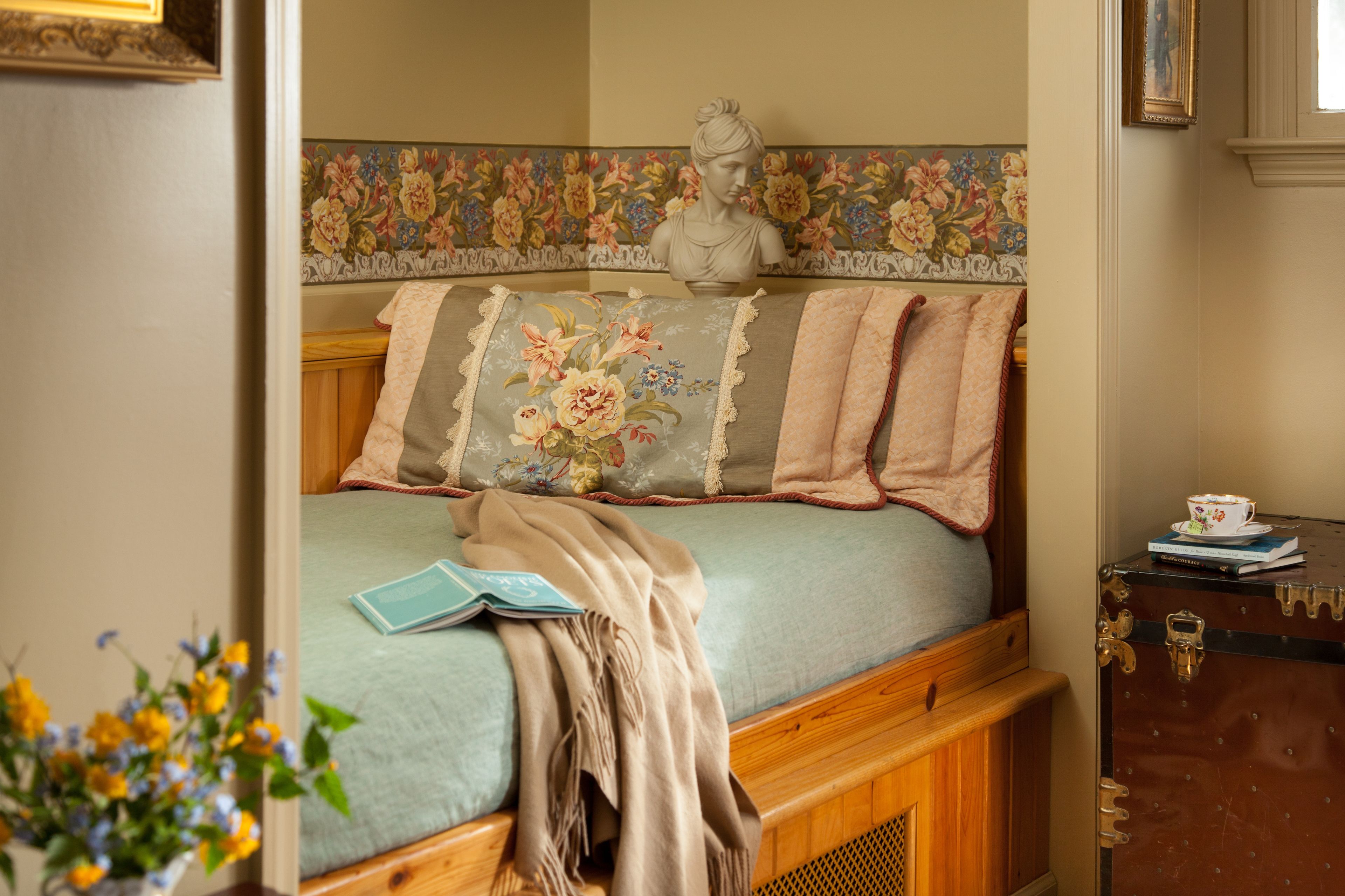 Un dormitorio con colcha estampada, cabecera de madera, una escultura de busto y una maleta antigua.