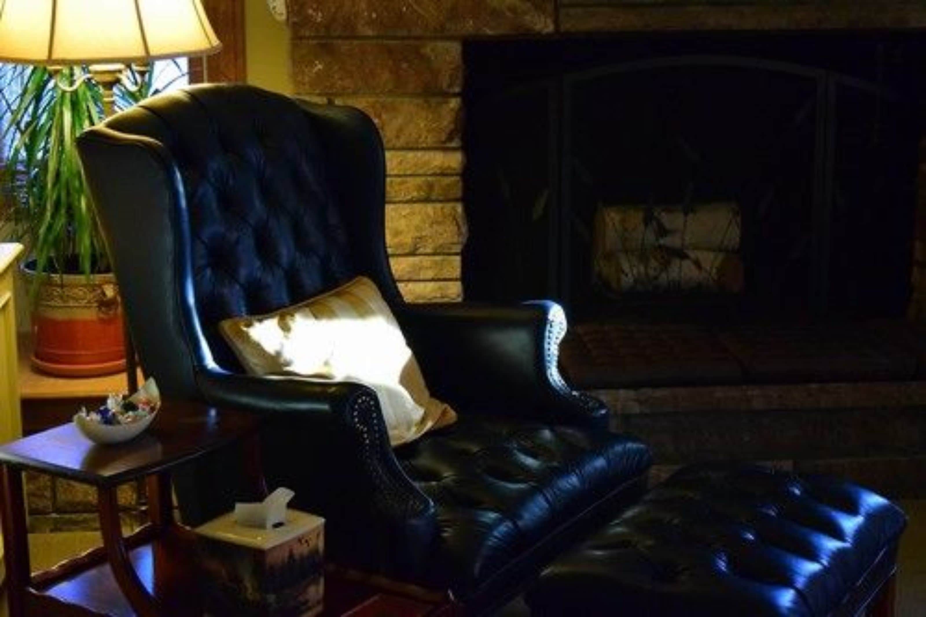 Un sillón de cuero oscuro con un cojín, una mesita con un bol y una caja de pañuelos, y una chimenea al fondo.