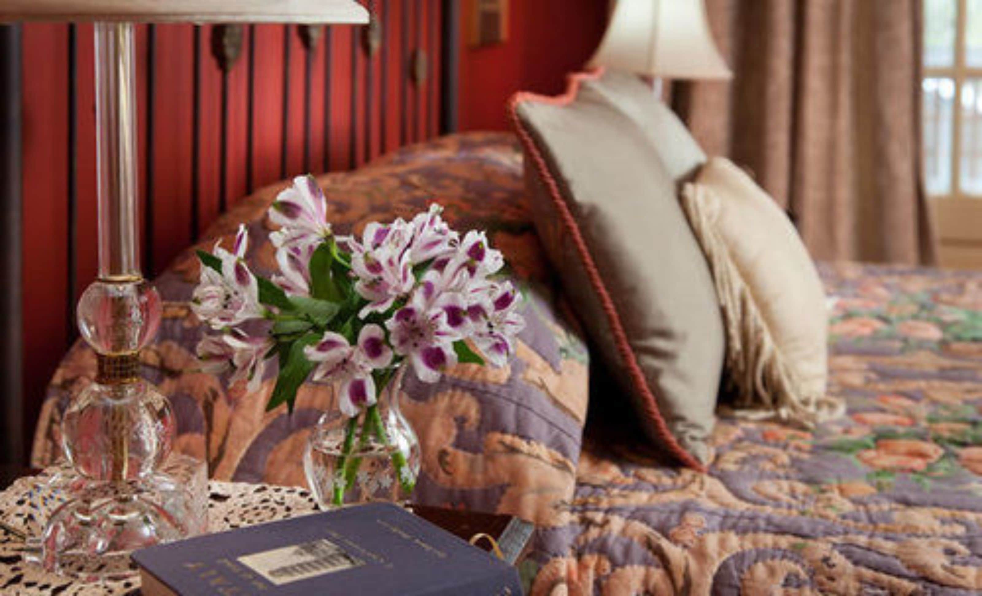 A neatly made bed with a floral-patterned bedspread, a vase with flowers, a book, and a lamp on a bedside table.