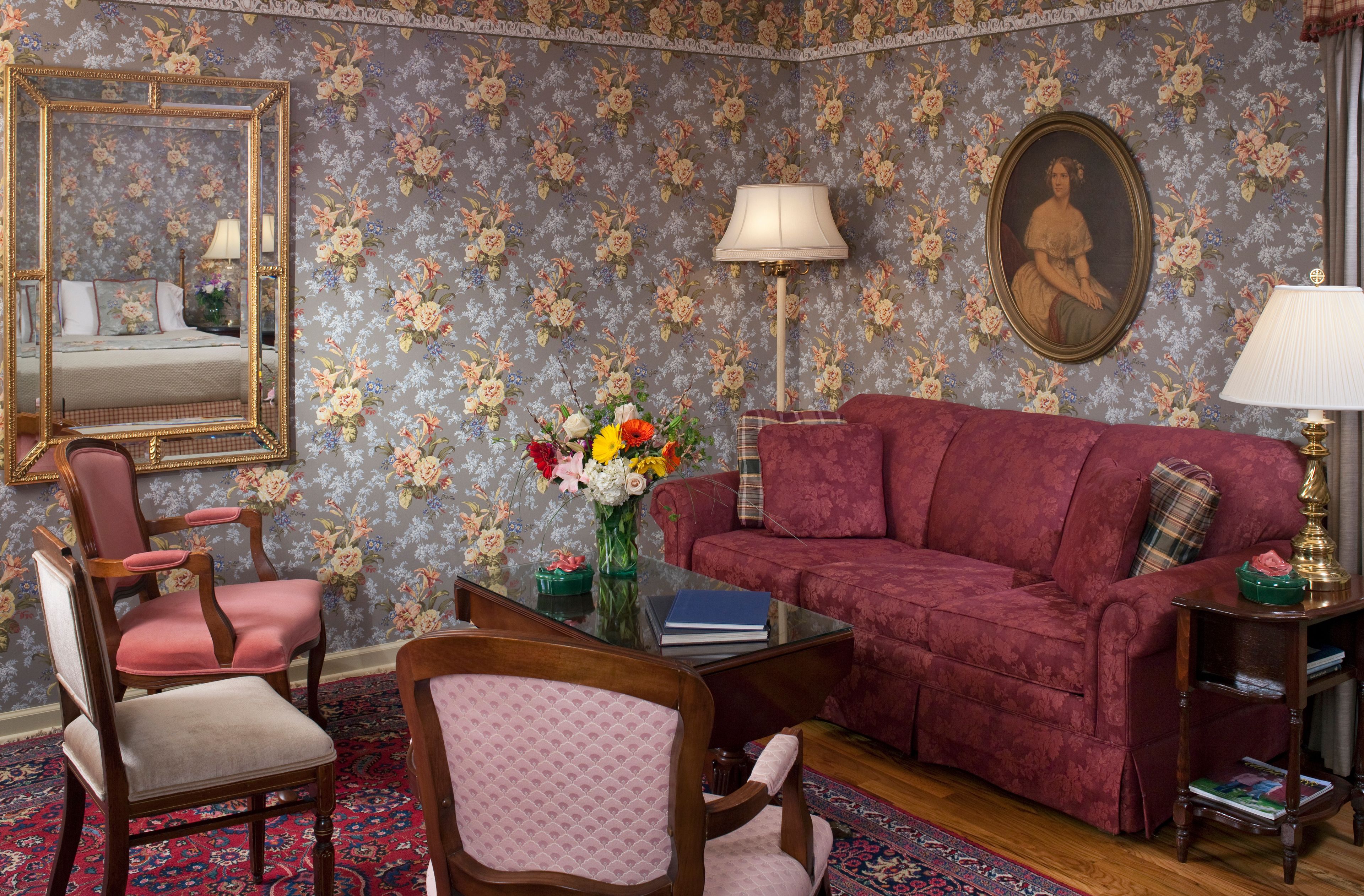 A room with a floral wallpaper, a red sofa, a wooden chair, a glass coffee table, and a mirror.