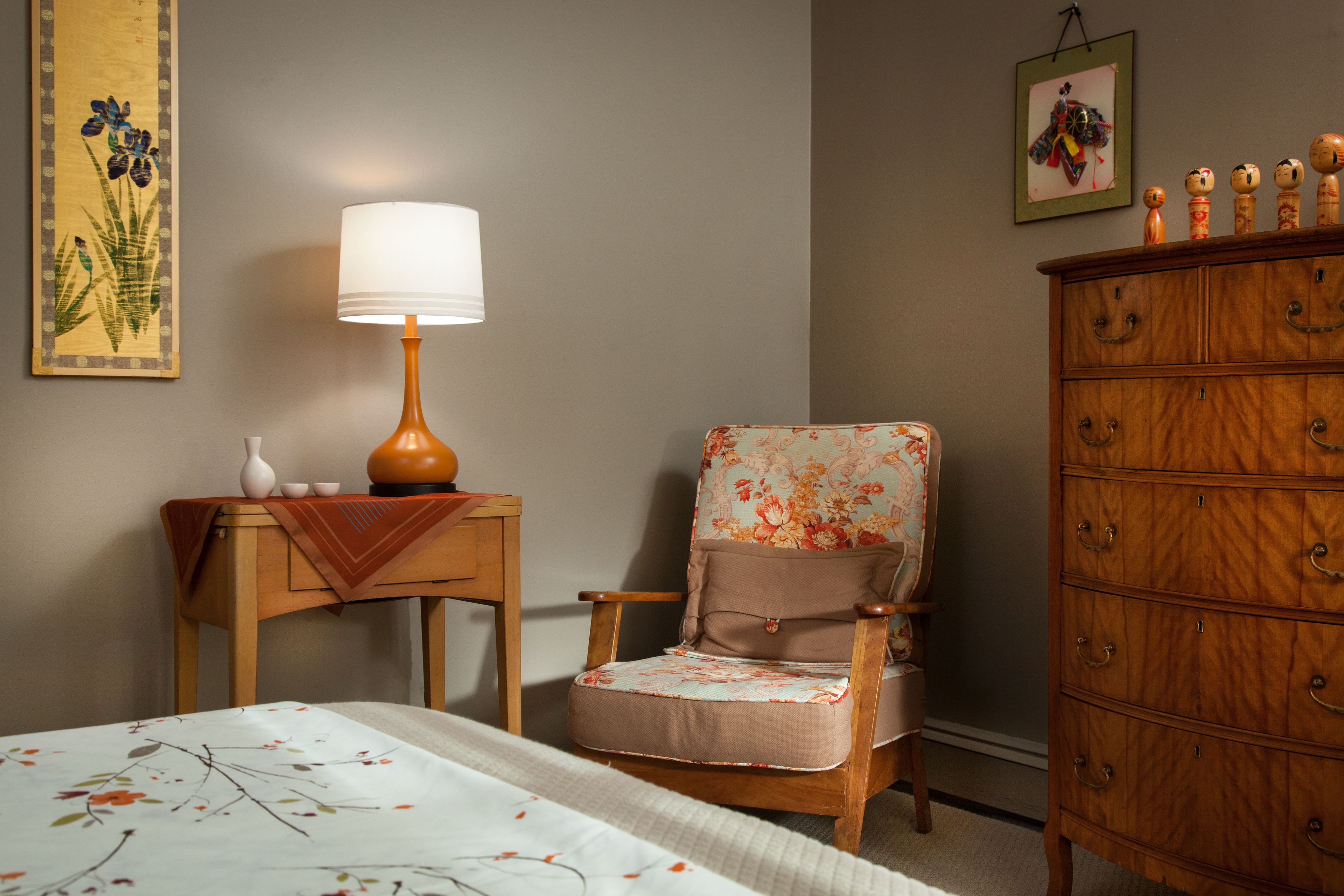 A room with a bed, a wooden chair with a floral cushion, a wooden dresser, a table with a lamp, and wall art.