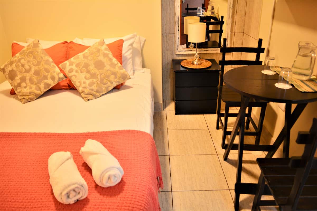 A hotel room with a bed, a red blanket, two rolled towels, a table with chairs, a lamp, and a pitcher of water.