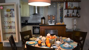 Un coin repas avec une table ronde dressée pour le petit-déjeuner, comprenant des assiettes, des tasses et un saladier de fruits. Des chaises sont disposées autour de la table, et on aperçoit en arrière-plan un espace cuisine avec des appareils électroménagers et des étagères.