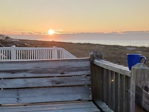 Eine Holzterrasse mit Geländer, die auf einen Strand und den Ozean im Sonnenuntergang blickt.