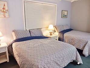 A bedroom with two beds, a nightstand with a lamp, a window with blinds, and a framed picture on the wall. - Westside- 3 Bedroom Home by RedAwning (Supply)