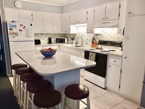 A kitchen with white cabinets, a microwave, a stove, and a refrigerator. There is a bar stool area with a bowl of fruit.
