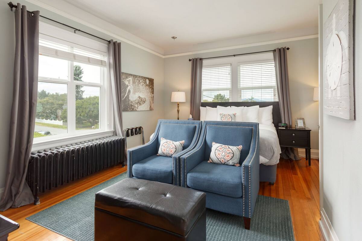 A bedroom with a large bed, a blue armchair, a wooden floor, a radiator, and a window with curtains.