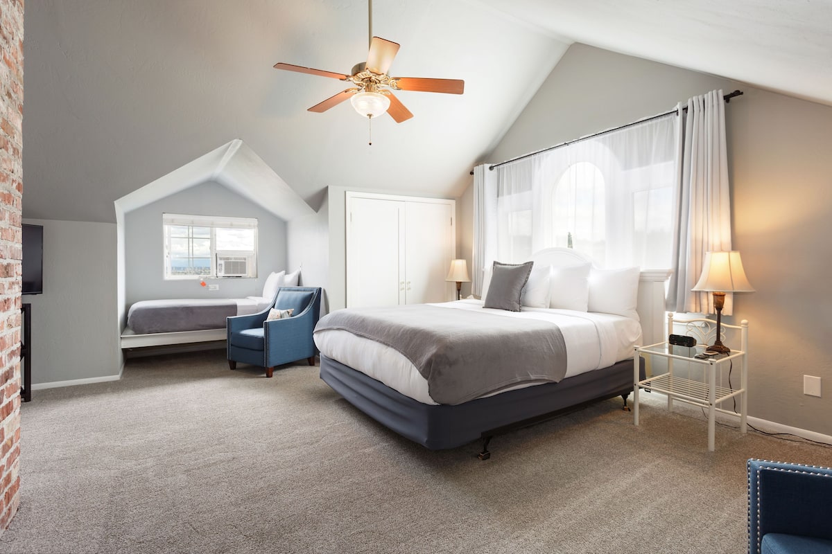 A bedroom with a bed, a chair, a desk with a lamp, a ceiling fan, and a window.