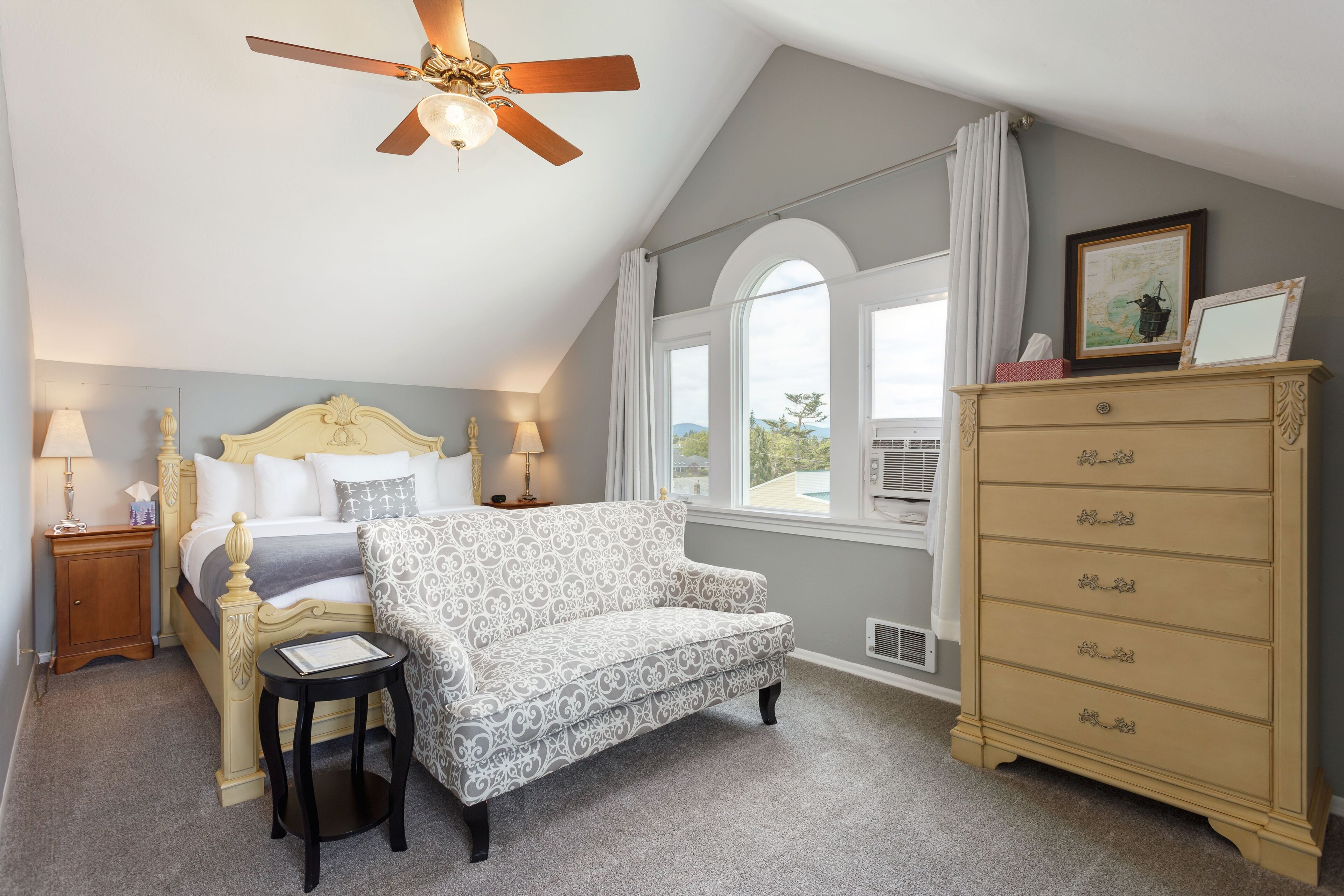 A bedroom with a bed, a patterned armchair, a dresser, a ceiling fan, and a window with a view.