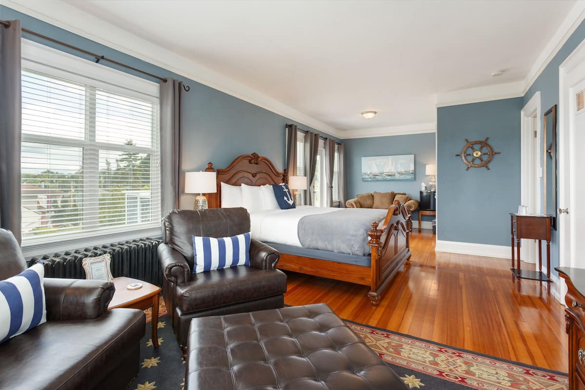A bedroom with a large bed, a leather armchair, a wooden floor, and a view of the outdoors through the window.