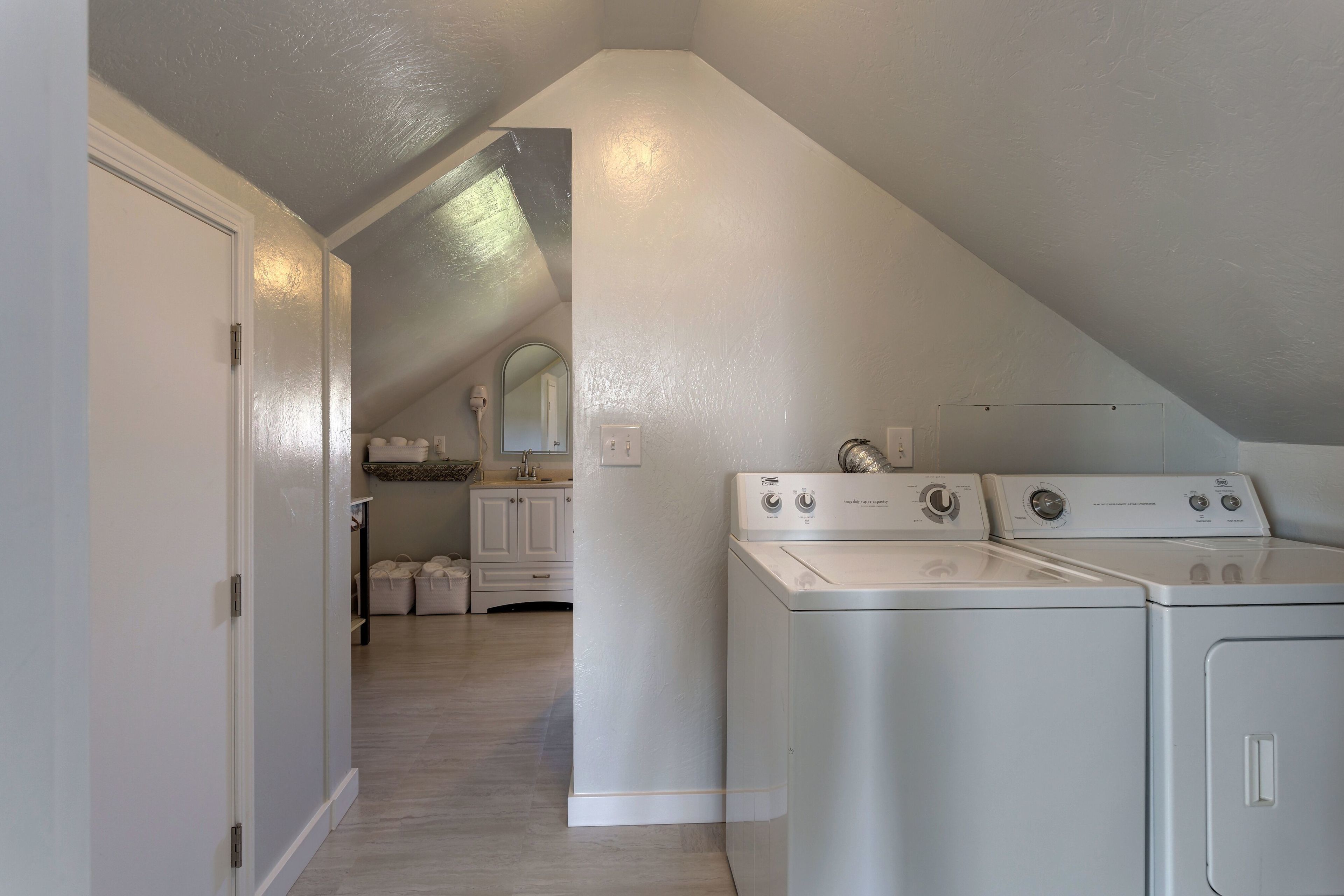 A laundry room with two front-loading washers and dryers, a white door, and a hallway leading to a bathroom with a vanity and cabinets.