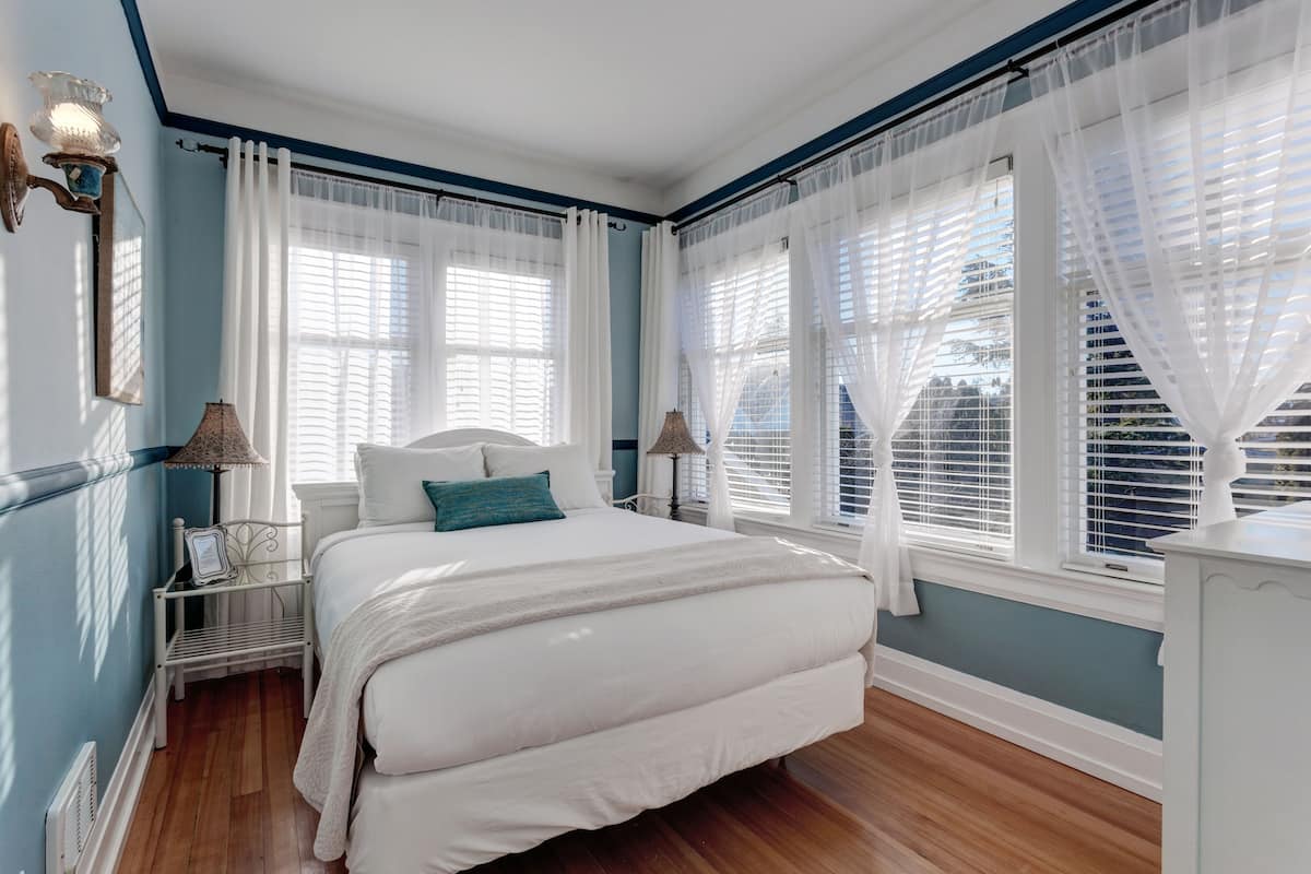 A bedroom with a large bed, white curtains, and a view of the city through the windows.