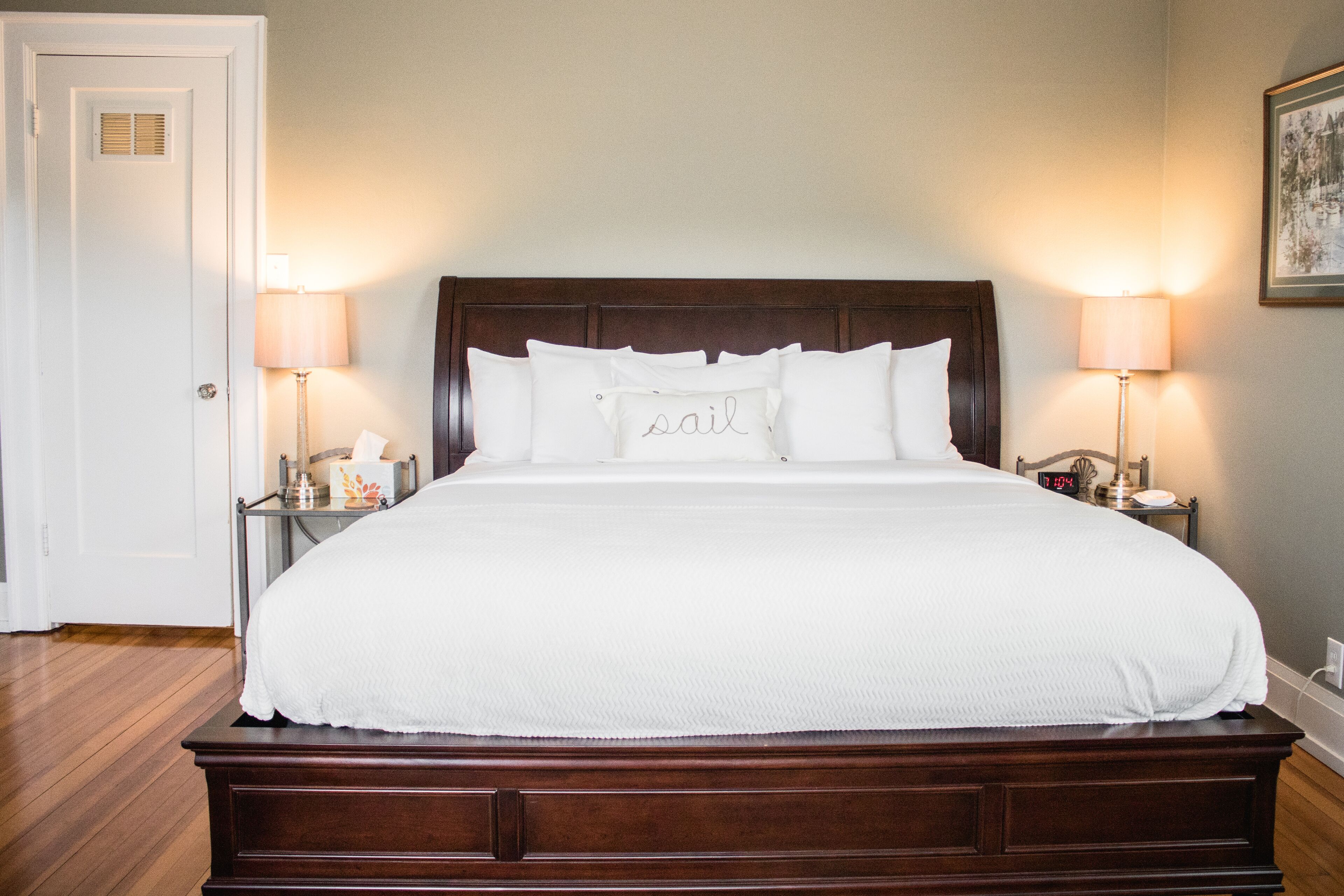A neatly made bed with a white comforter and pillows, two bedside lamps, a nightstand with a lamp, and a framed picture on the wall.