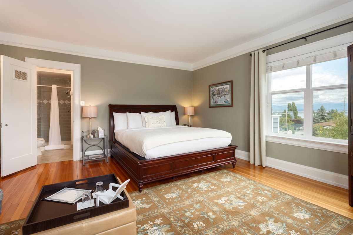 A bedroom with a large bed, a wooden floor, a view through a window, and a bathroom visible through an open door.