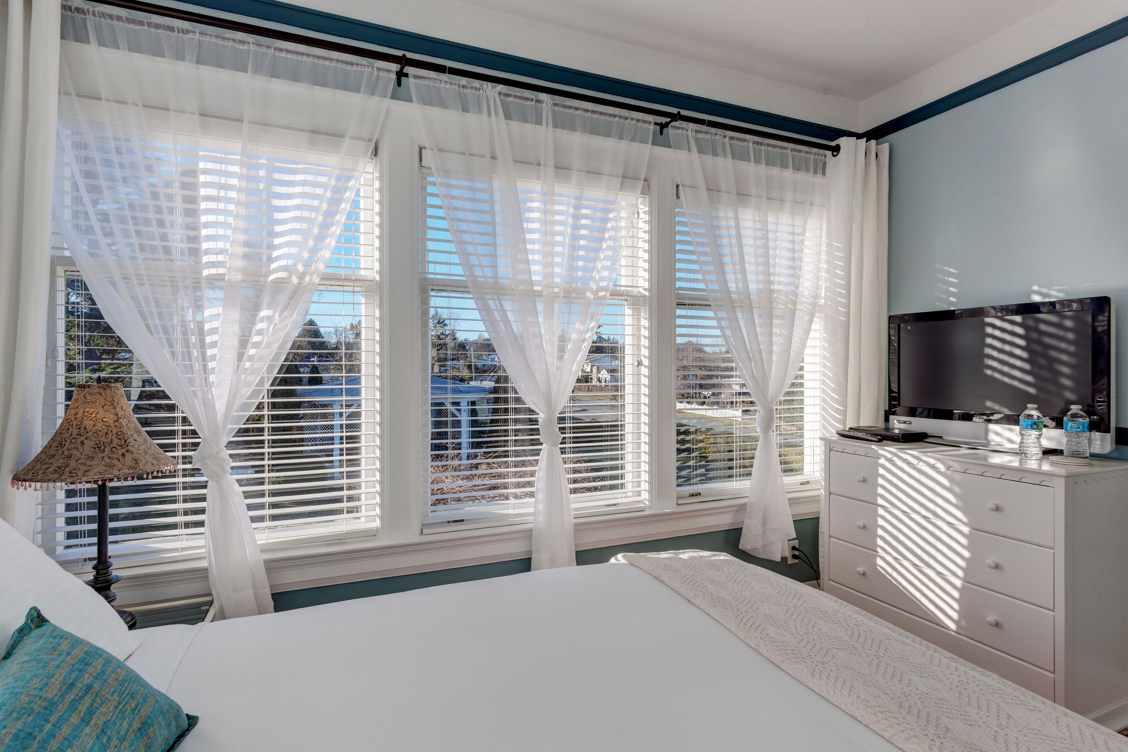 A bedroom with a bed, a TV, a dresser, and a window with sheer curtains.