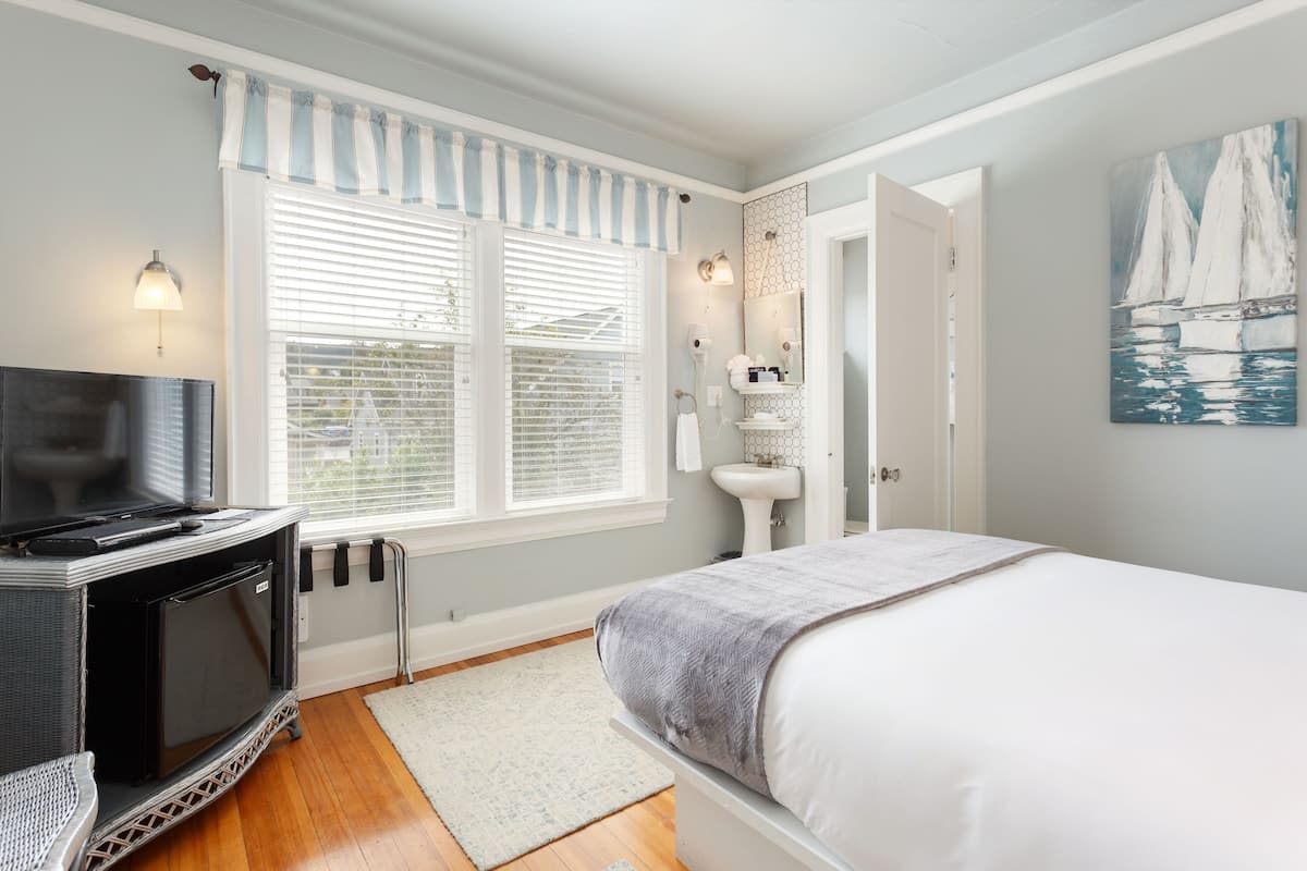 A bedroom with a TV, a radiator, a window with blinds, a bedside table, and a painting on the wall.
