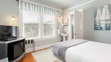 A bedroom with a bed, a TV, a radiator, a window with blinds, a sink, and a painting on the wall.