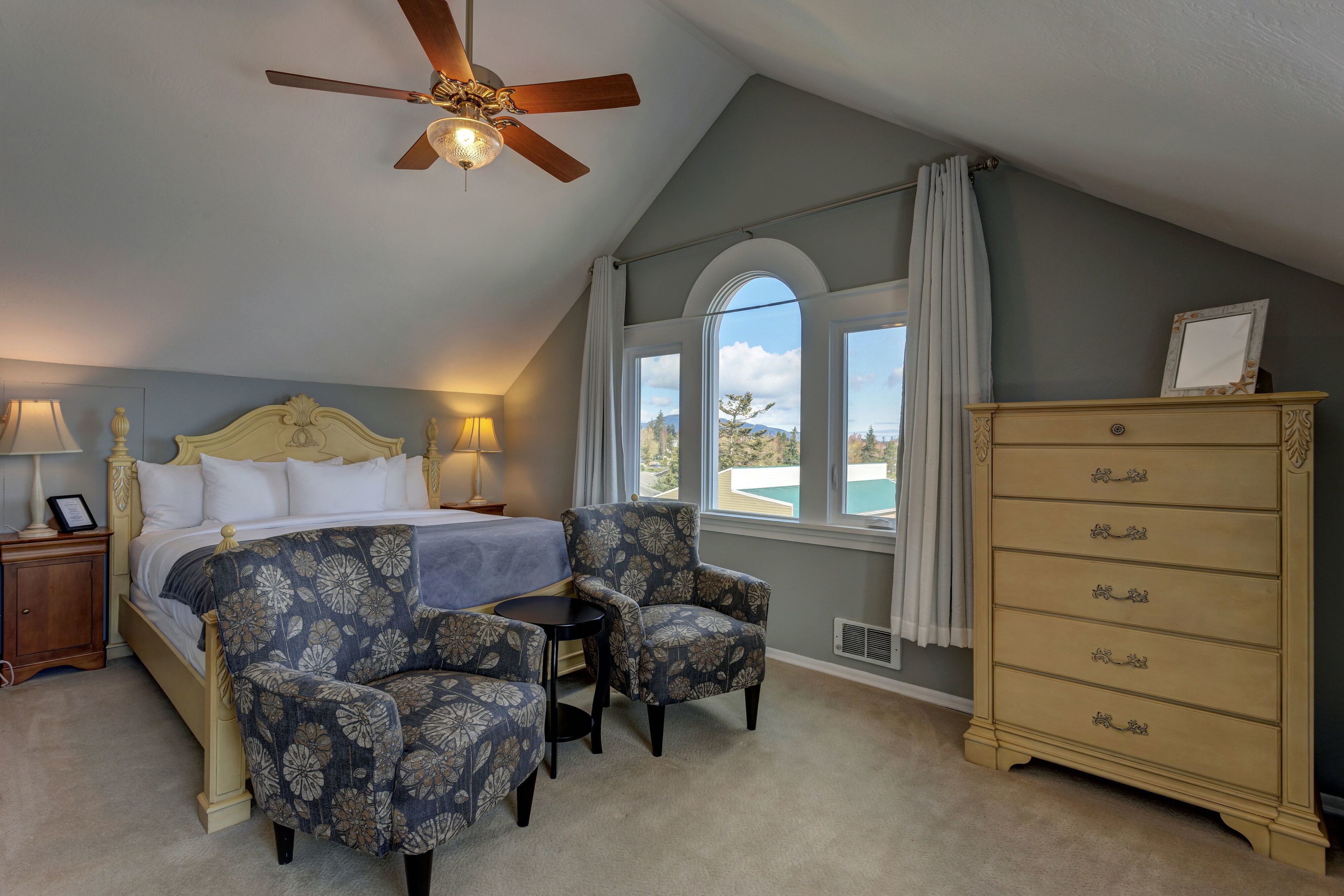 A bedroom with a bed, two armchairs, a dresser, a ceiling fan, and a view of the outdoors.