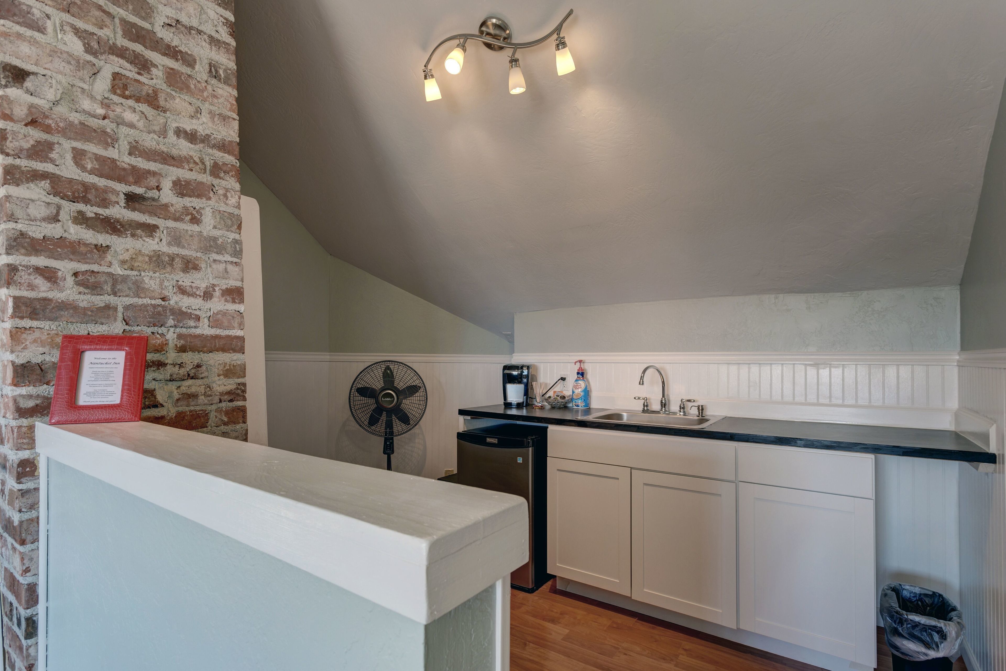 Une cuisine avec un mur en briques, des meubles blancs, un plan de travail noir, un évier, un ventilateur et un luminaire au plafond.