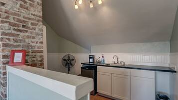 Une cuisine avec un mur en briques, des meubles blancs, un plan de travail noir, un évier, un ventilateur et un luminaire au plafond.