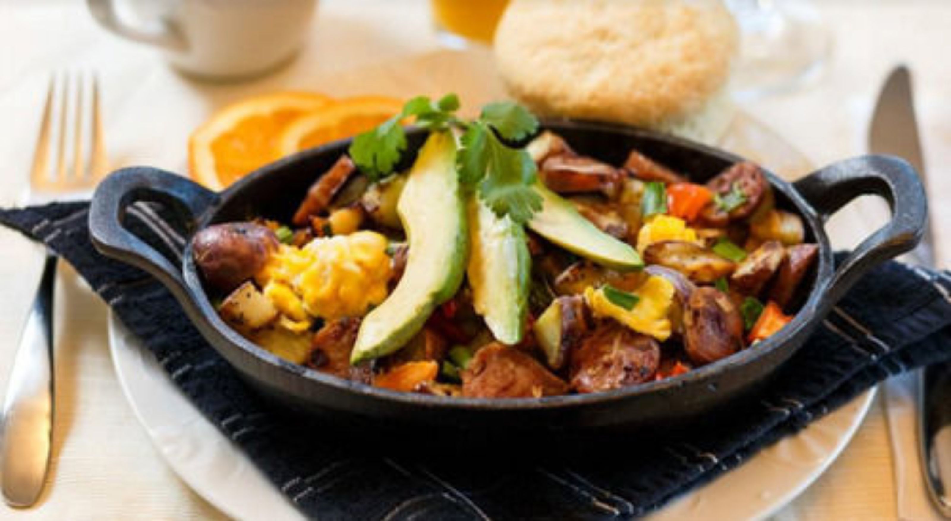 A skillet with a breakfast dish, including eggs, potatoes, and avocado, on a white plate with a black napkin underneath.