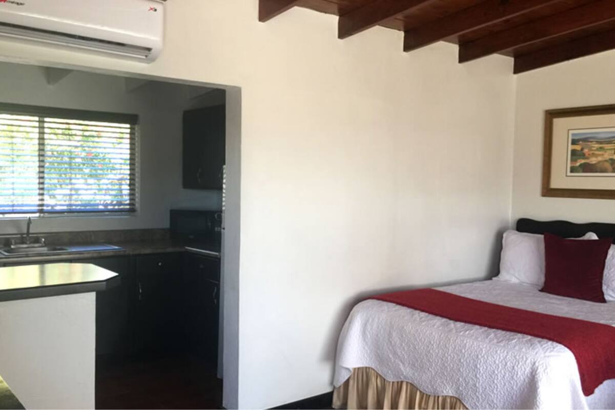 Ein Schlafzimmer mit Bett, einem Fenster mit Jalousien, einer Küchenzeile und einem Deckenventilator.