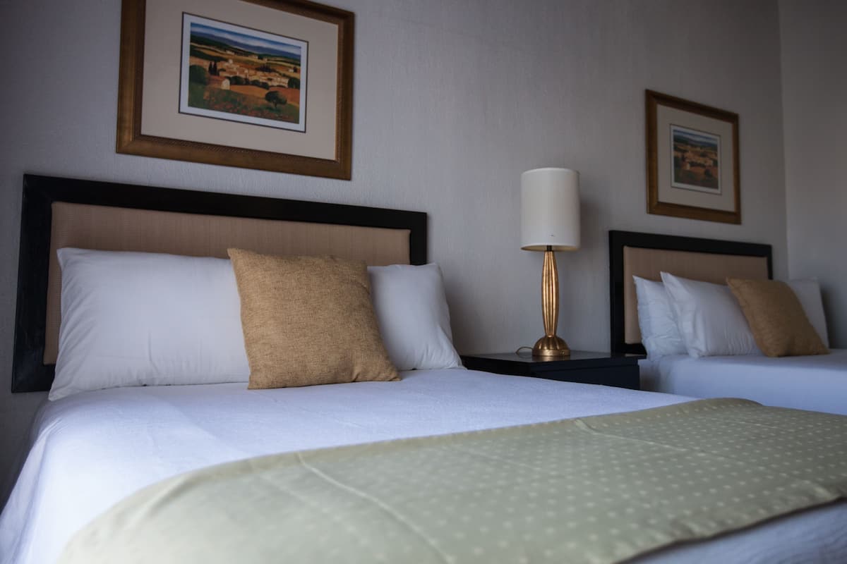 Ein Hotelzimmer mit zwei Betten, jeweils mit Kopfteil, Nachttischlampe und einem gerahmten Bild an der Wand.