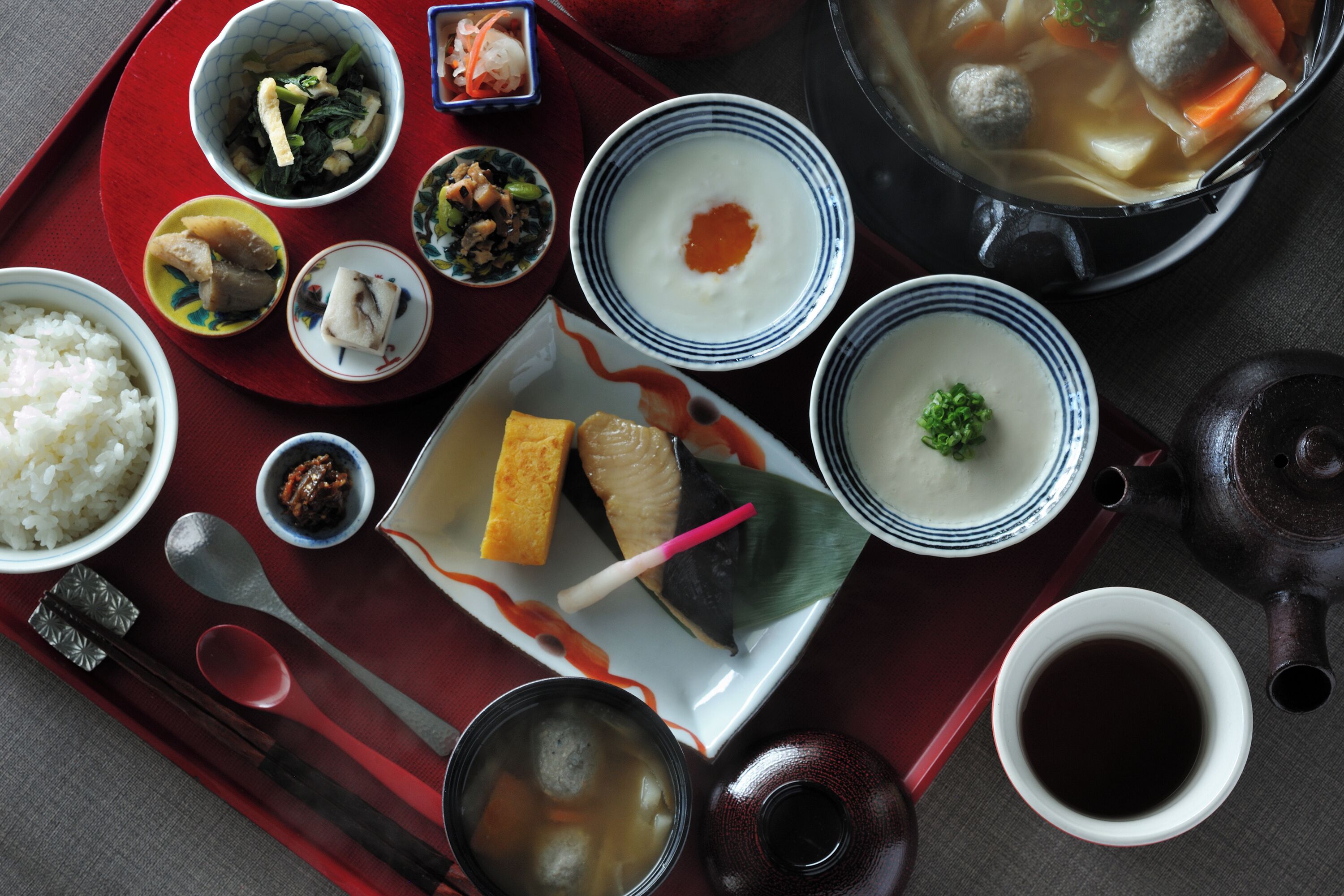 A variety of Japanese dishes including rice, soup, and meatballs, served on a red tray with chopsticks and a teapot.