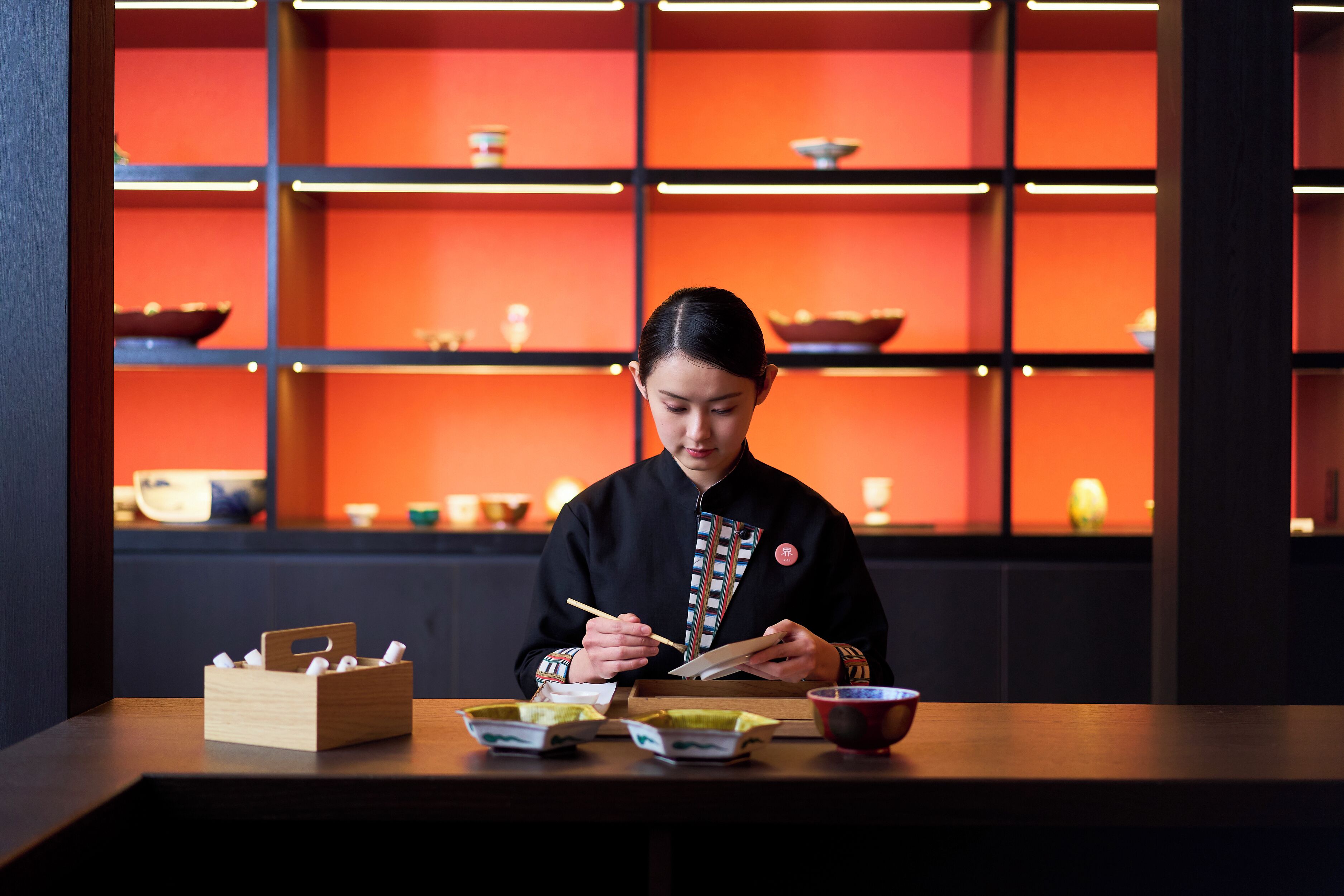 A person dining at a table with chopsticks and bowls, in a room with shelves in the background.