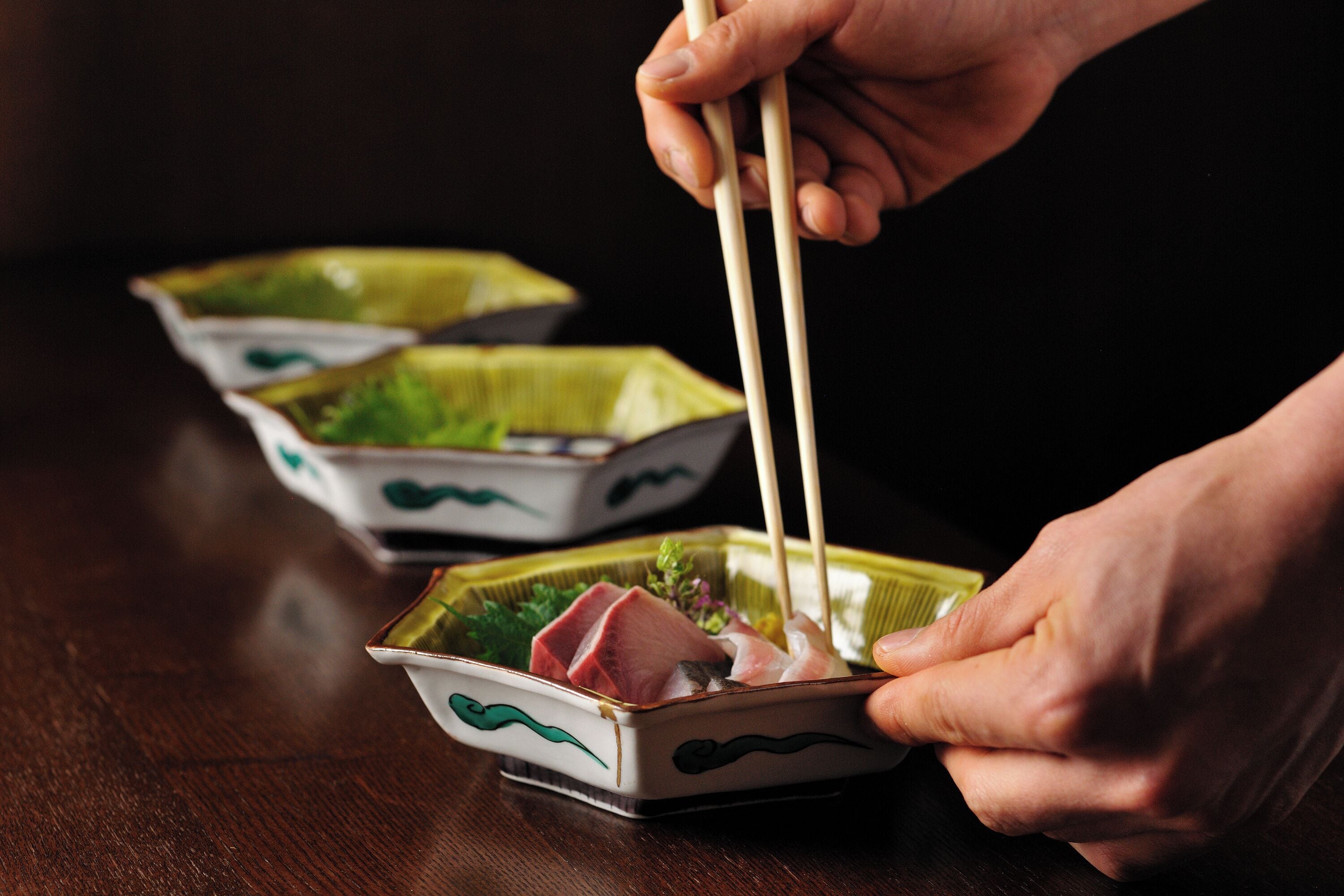 A person holding chopsticks over a small dish with food, likely sushi, on a wooden table.